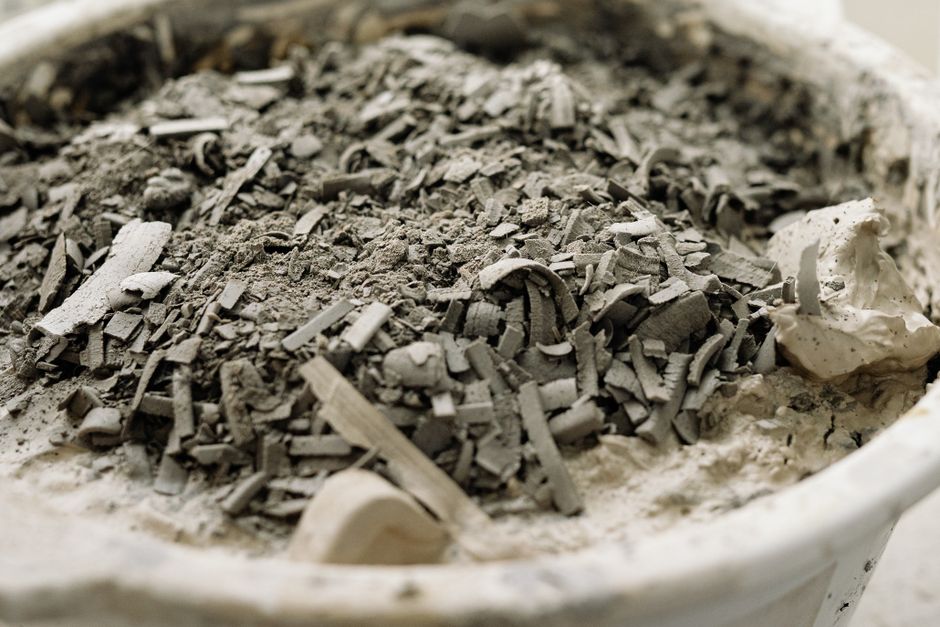 Close-up image of grey ash and small chippings of burnt wood and charcoal in a white ceramic or plastic container, with a rough, uneven texture on the ash surface. The ash appears fine with some large